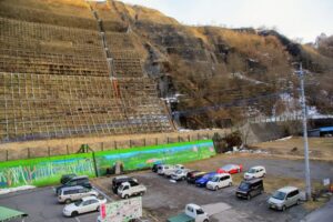 駅前駐車場と法面工事をした壁面。駐車場の端に竣工記念碑あり。壁面に施された排水溝(タテのスジ)の下の方が赤くなっているのは、鉱水の影響とのこと、地元の人のお話。